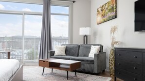 Een moderne slaapkamer met een groot raam, een grijze bank, een houten salontafel, een zwarte dressoir en een schilderij aan de muur.