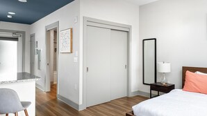 Een moderne hotelkamer met een keukenhoek, een grote inloopkast, een nachtkastje met een lamp en een bed met een perzikkleurig kussen.