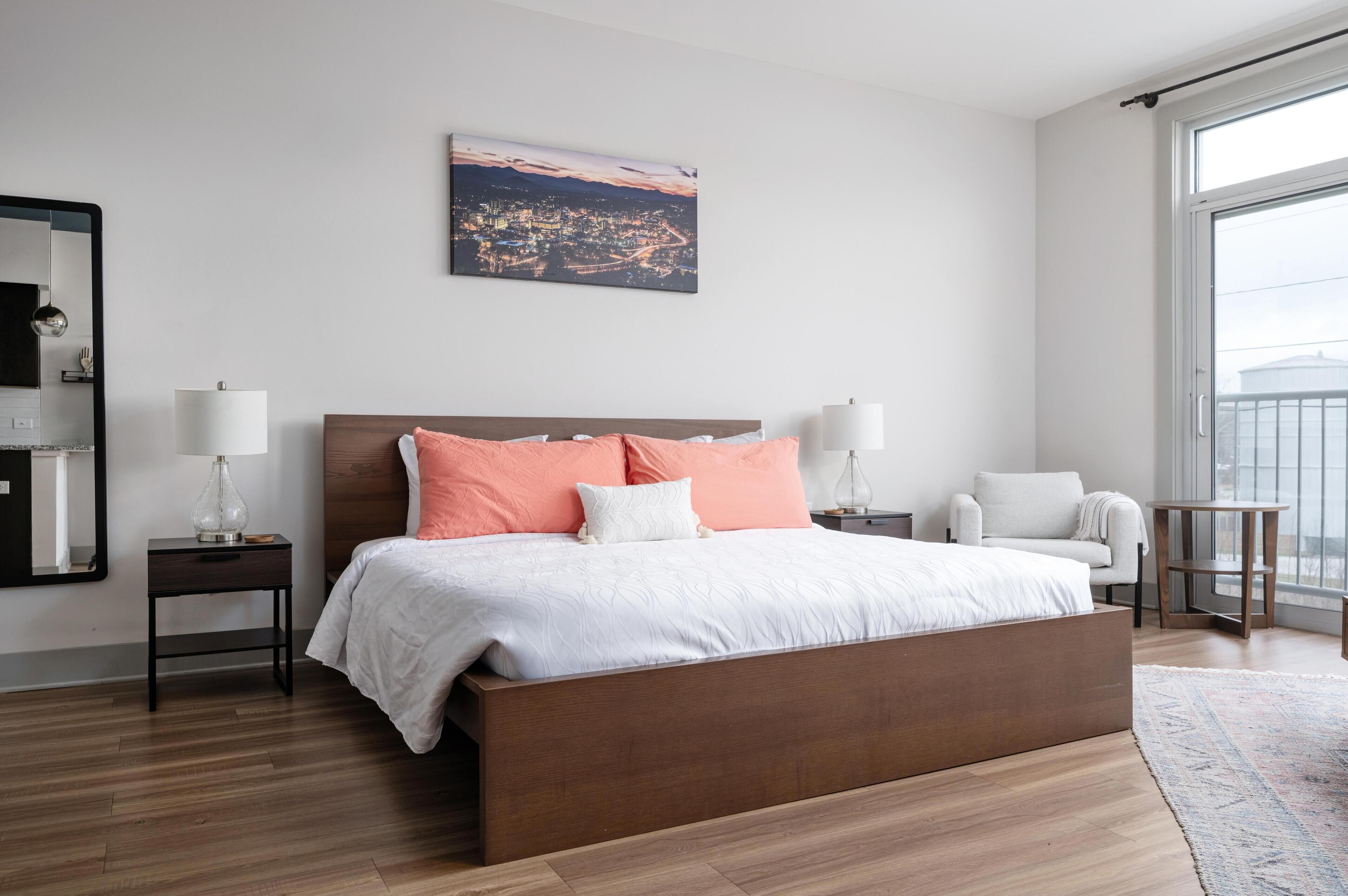 A modern bedroom with a large bed, two bedside tables with lamps, a chair, and a cityscape view.