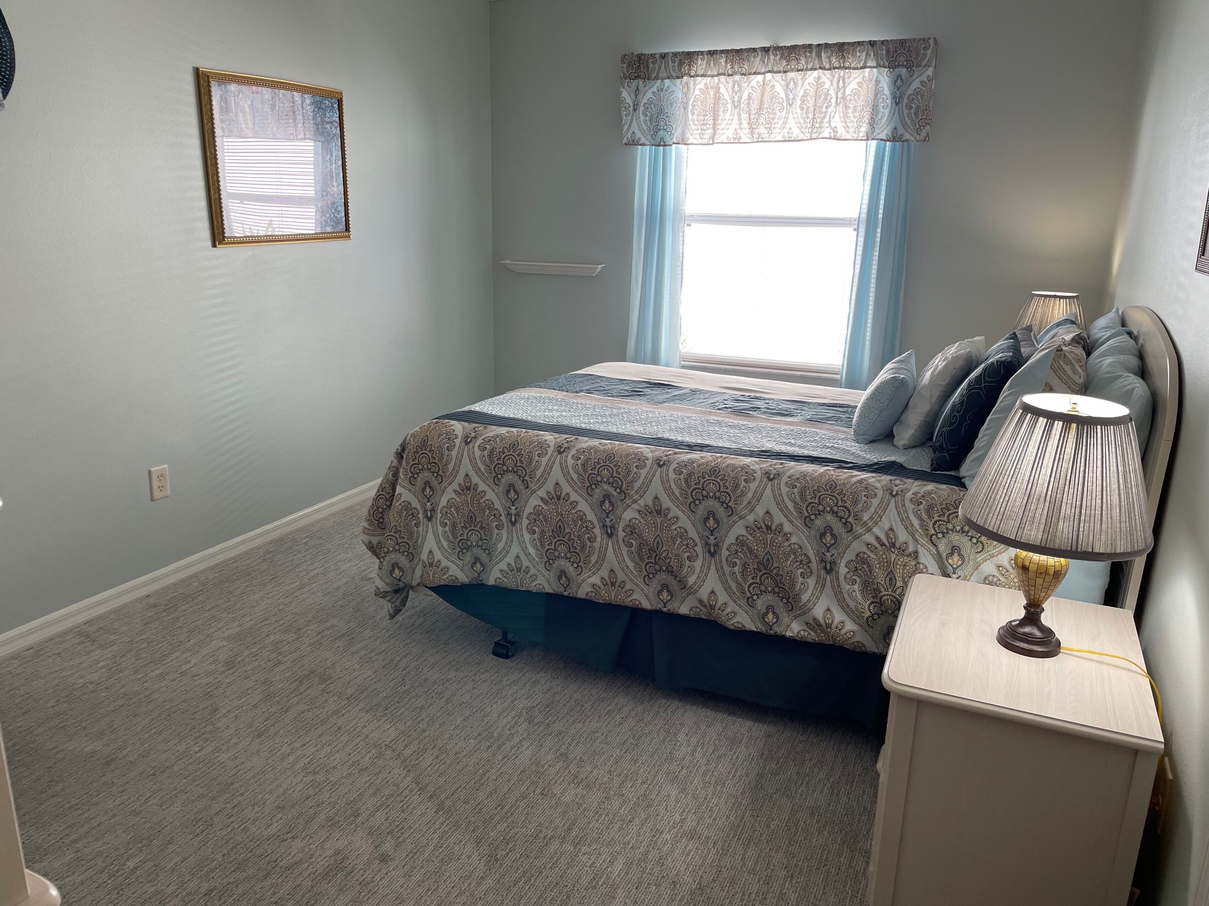 A bedroom with a bed, a nightstand, a lamp, a window with curtains, and a wall-mounted picture frame.