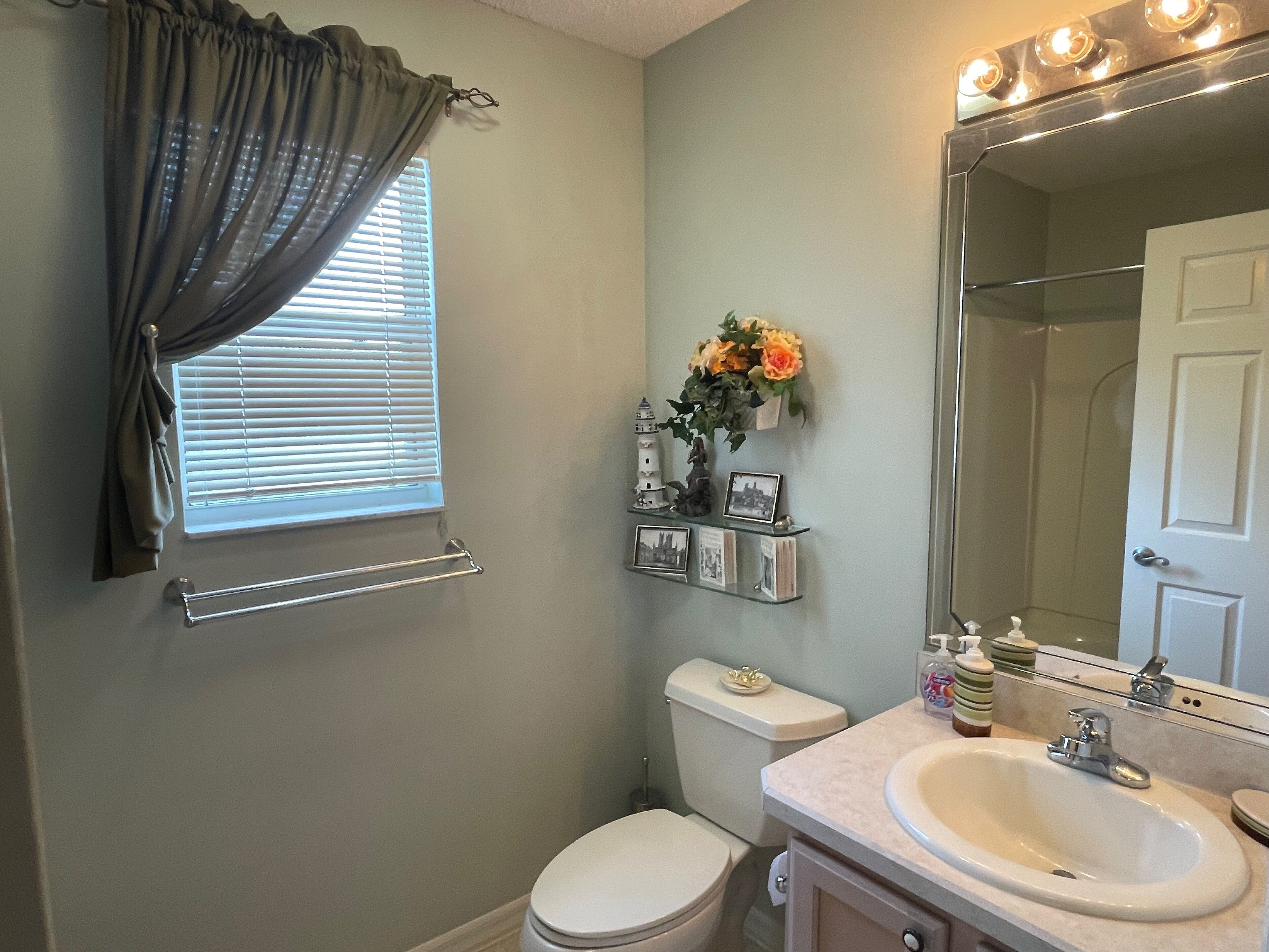 A bathroom with a toilet, sink, vanity, and a shower. There is a window with blinds, a towel rack, and a mirror with lights above the sink.