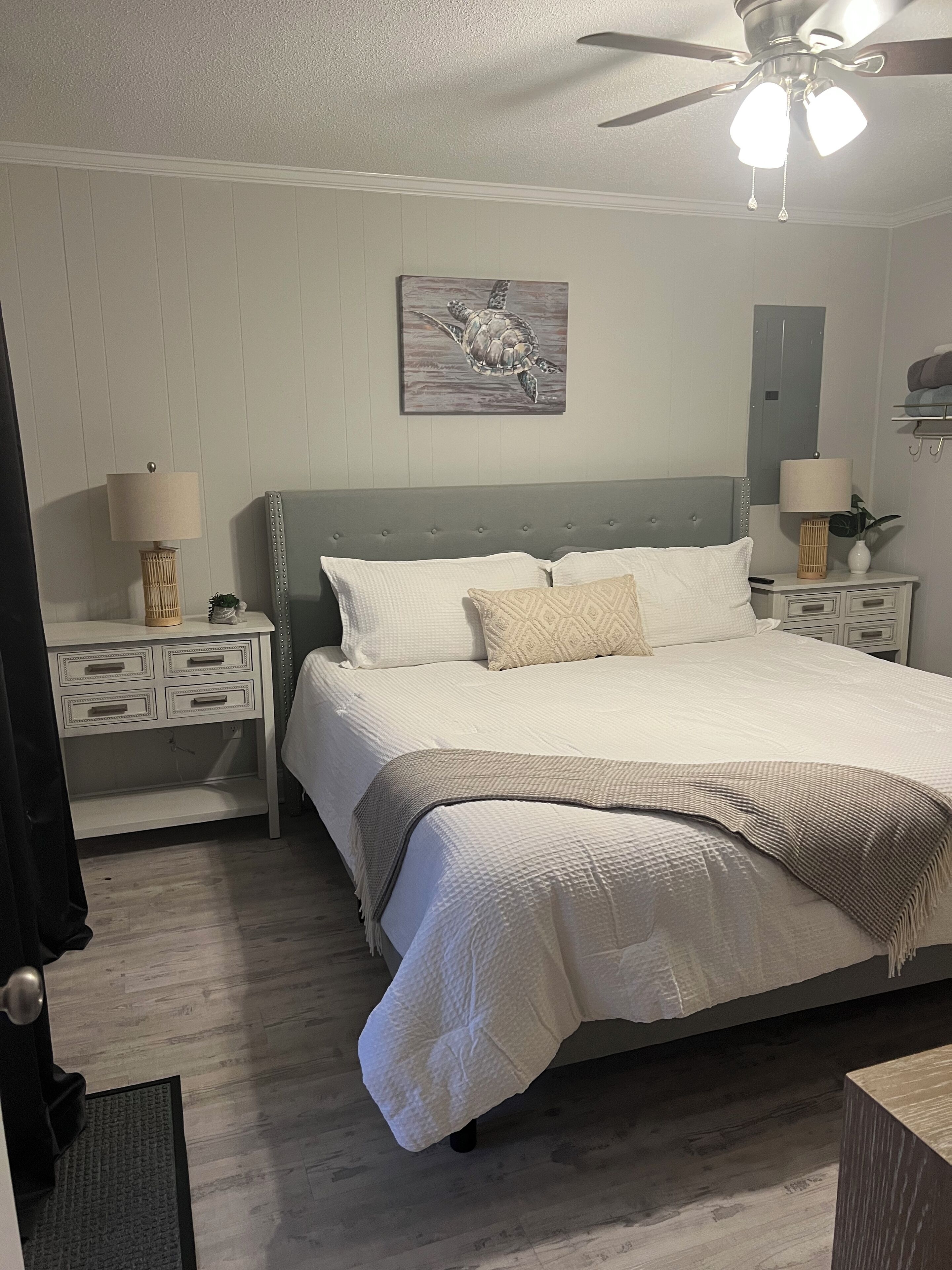 A bedroom with a bed, nightstands, a ceiling fan, a wall-mounted painting, and a door.