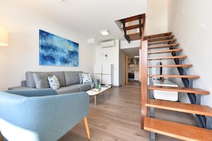 A modern living room with a grey sofa, a blue armchair, and a coffee table. A wooden staircase leads to an upper level.