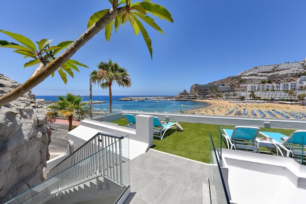 A balcony with a view of a beach and ocean, a palm tree, and a staircase.