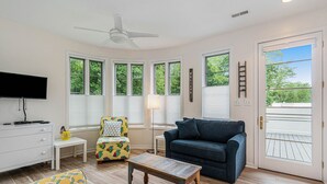 Un salon comprenant un téléviseur, un canapé, un fauteuil, une petite table et une fenêtre donnant sur des arbres.
