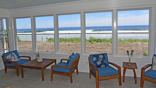 A room with large windows overlooking a beach, wooden furniture, and a small table with a vase. - Luxury Oceanfront 5 Bedroom 3.5 Bathroom Private Parsons Beachfront Paradise (Kennebunk)