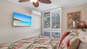 A bedroom with a bed, a ceiling fan, a window with blinds, a wall-mounted TV, and a framed picture.