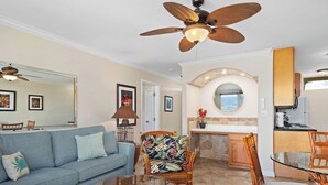 A living room with a sofa, chairs, a coffee table, a ceiling fan, and a kitchen area with a counter and cabinets.