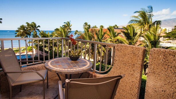 A balcony with a table and chairs, overlooking a pool and palm trees, with a view of the ocean and mountains in the background.