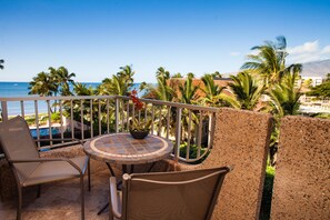 A balcony with a table and chairs, overlooking a pool and palm trees, with a view of the ocean and mountains in the background.