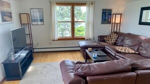 A living room with a brown leather sofa, a TV on a stand, a window with curtains, and a framed picture on the wall. - Location!! Ocean views. Easy walk to beaches, restaurants, shops. Quiet street.  (Middletown)