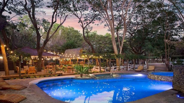 Un espace piscine doté d’un sol en pierre, entouré d’arbres, et un coin repas agrémenté de guirlandes lumineuses.