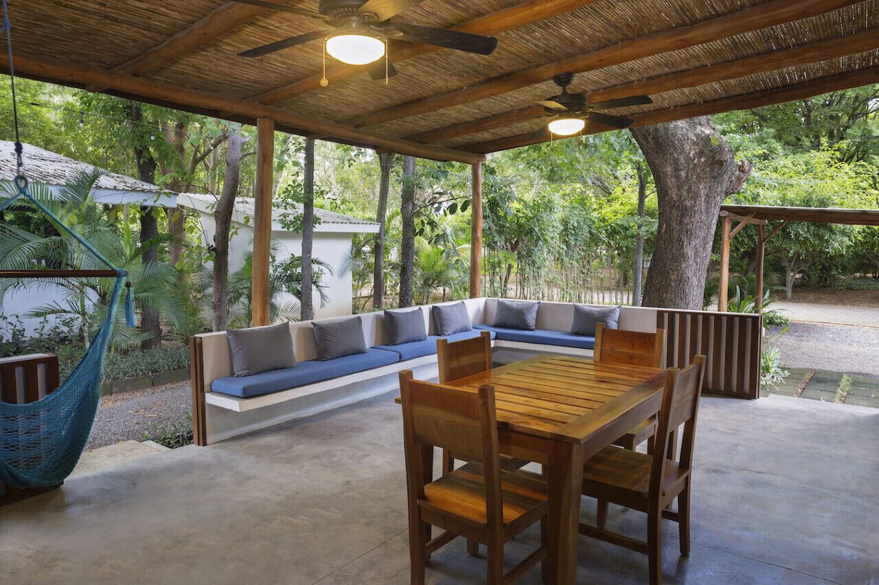 A covered outdoor seating area with a wooden table and chairs, surrounded by lush greenery.