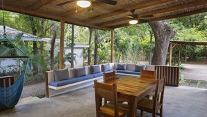Un espace extérieur aménagé avec une zone de repos couverte, comprenant une table et des chaises en bois, le tout entouré d’une végétation luxuriante.