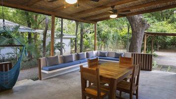 A covered outdoor seating area with a wooden table and chairs, surrounded by lush greenery.