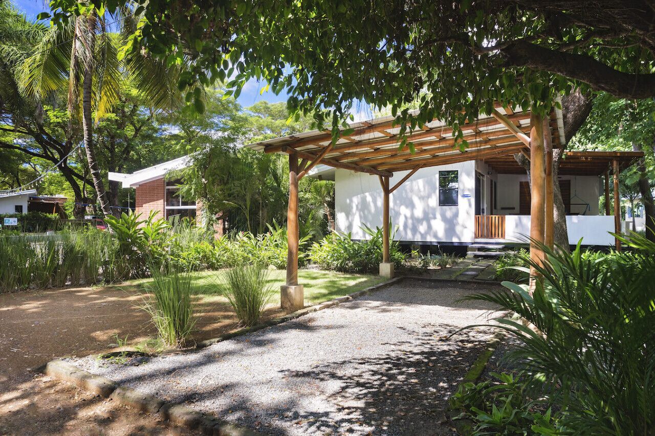 A covered outdoor area with a wooden pergola, surrounded by lush greenery and a gravel path.