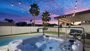 A hot tub with a control panel, surrounded by a pool, under a pergola with string lights, palm trees, and a sunset sky. - HOT TUB • HEATED POOL • Close to Dining & Shopping (Scottsdale)