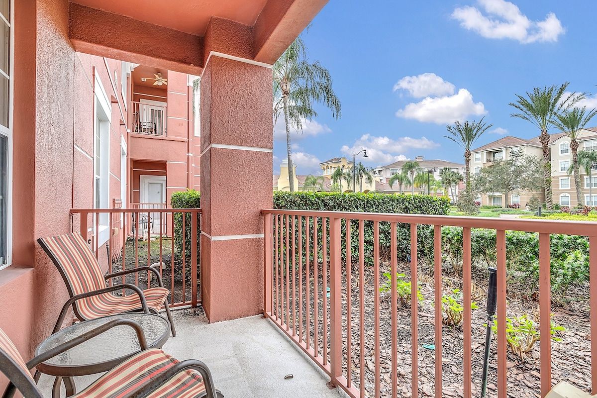 A balcony with chairs, a railing, and a view of neighboring buildings and palm trees.