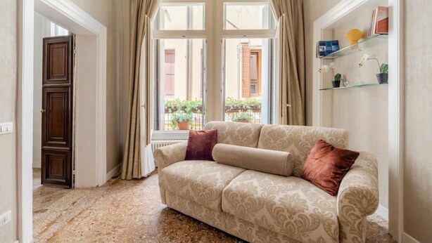A living room with a beige patterned sofa, a brown cushion, a glass shelf with books and decorative items, and a view through a window with curtains. - Charming apartment not far from Piazza San Marco (Venezia)
