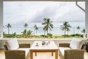 A balcony with wicker furniture, a table, and a view of the ocean and palm trees. - The Oyster Bay Hotel Suites (Dar es Salaam)