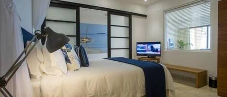 Une chambre à coucher comprenant un lit, un bureau, une chaise, un téléviseur et une fenêtre avec des stores.