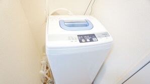 Een compacte, witte wasmachine met een bedieningspaneel en een blauwe hendel, geplaatst in een hoek met een houten vloer en een witte muur.