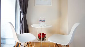 Een kleine kamer met een ronde tafel, twee witte stoelen, een ingelijst schilderij aan de muur en een raam met gordijnen.