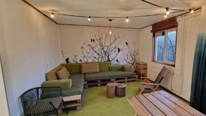 A cozy room with a green sofa, a wooden table, and a chair. There is a window with curtains, a radiator, and a wall with a tree mural.