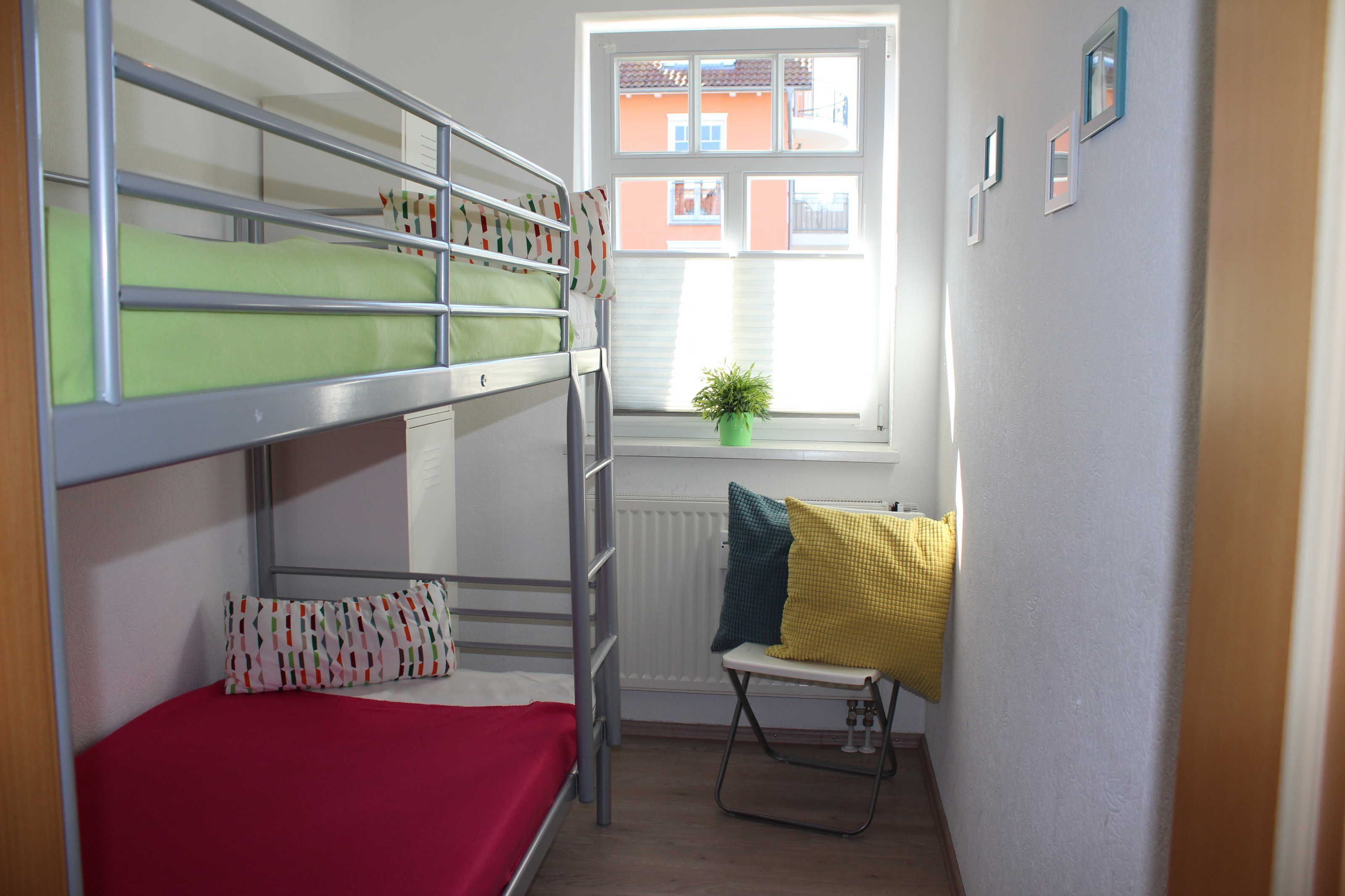 A bunk bed room with a red bedspread, a small table with a chair, and a window with a plant.