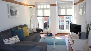 A living room with a grey sofa, a white TV stand, and a large window with blinds. - (311/1) 3-room apartment - apartment house Strandburg (Kühlungsborn)