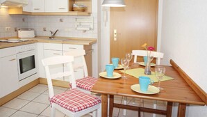 A compact kitchen with white cabinets, a built-in oven, and a dining area with a wooden table, chairs, and a hanging light fixture. - (311/1) 3-room apartment - apartment house Strandburg (Kühlungsborn)