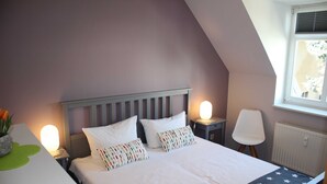 A bedroom with a sloped ceiling, a bed with white bedding and colorful pillows, a bedside table with a lamp, a chair, and a window with blinds. - (311/1) 3-room apartment - apartment house Strandburg (Kühlungsborn)