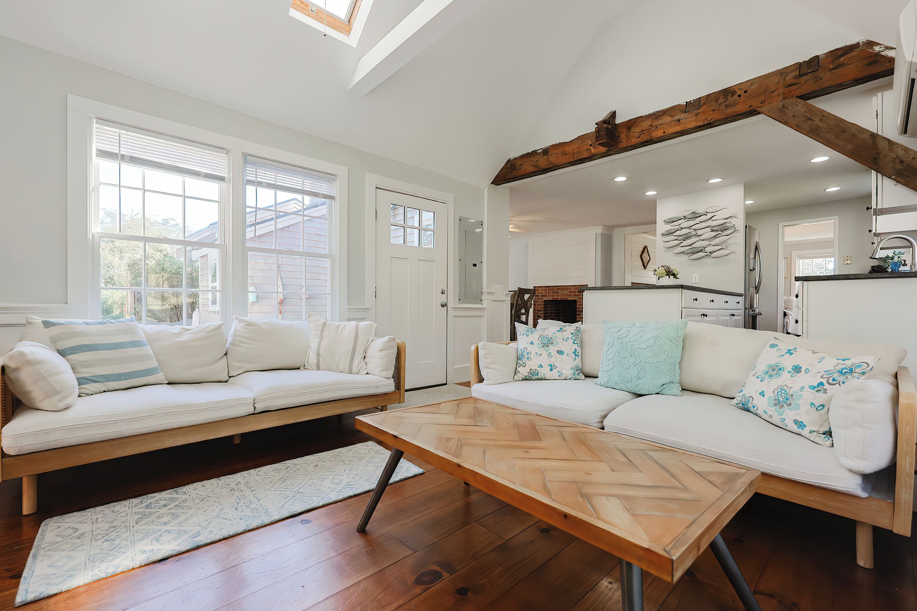 A spacious living room with a wooden coffee table, two white sofas with patterned cushions, a herringbone-patterned rug, and a view of the kitchen through an open door.