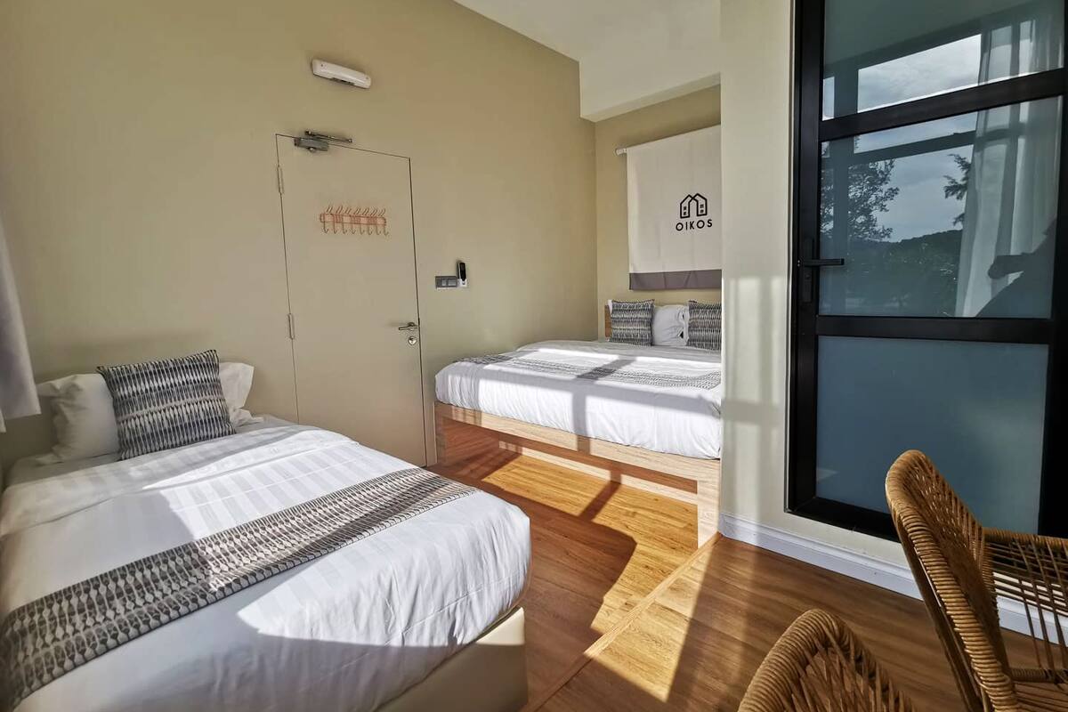 A hotel room with two beds, a large window, a wicker chair, and a towel rack.
