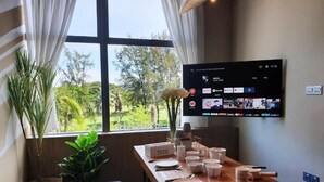 A room with a wooden table set for dining, a flat-screen TV mounted on the wall displaying a streaming service interface, and a view of greenery outside through large windows. - Oikos Poshtel (Kota Kinabalu)