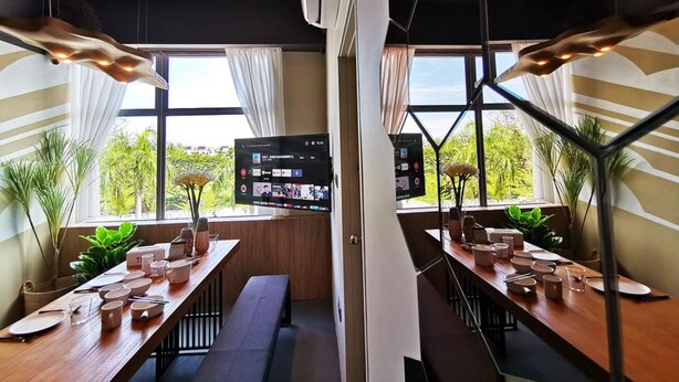 A modern conference room with a large screen displaying a presentation, wooden tables set for a meeting, and large windows with a view of greenery outside. - Oikos Poshtel (Kota Kinabalu)