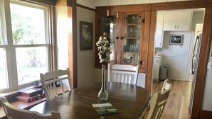 A dining area with a wooden table, white chairs, a vase with flowers, and a view of the outdoors through a large window. - 4 bedroom, 116 acre Farmhouse 10 minutes from downtown  Charlevoix  (Charlevoix)