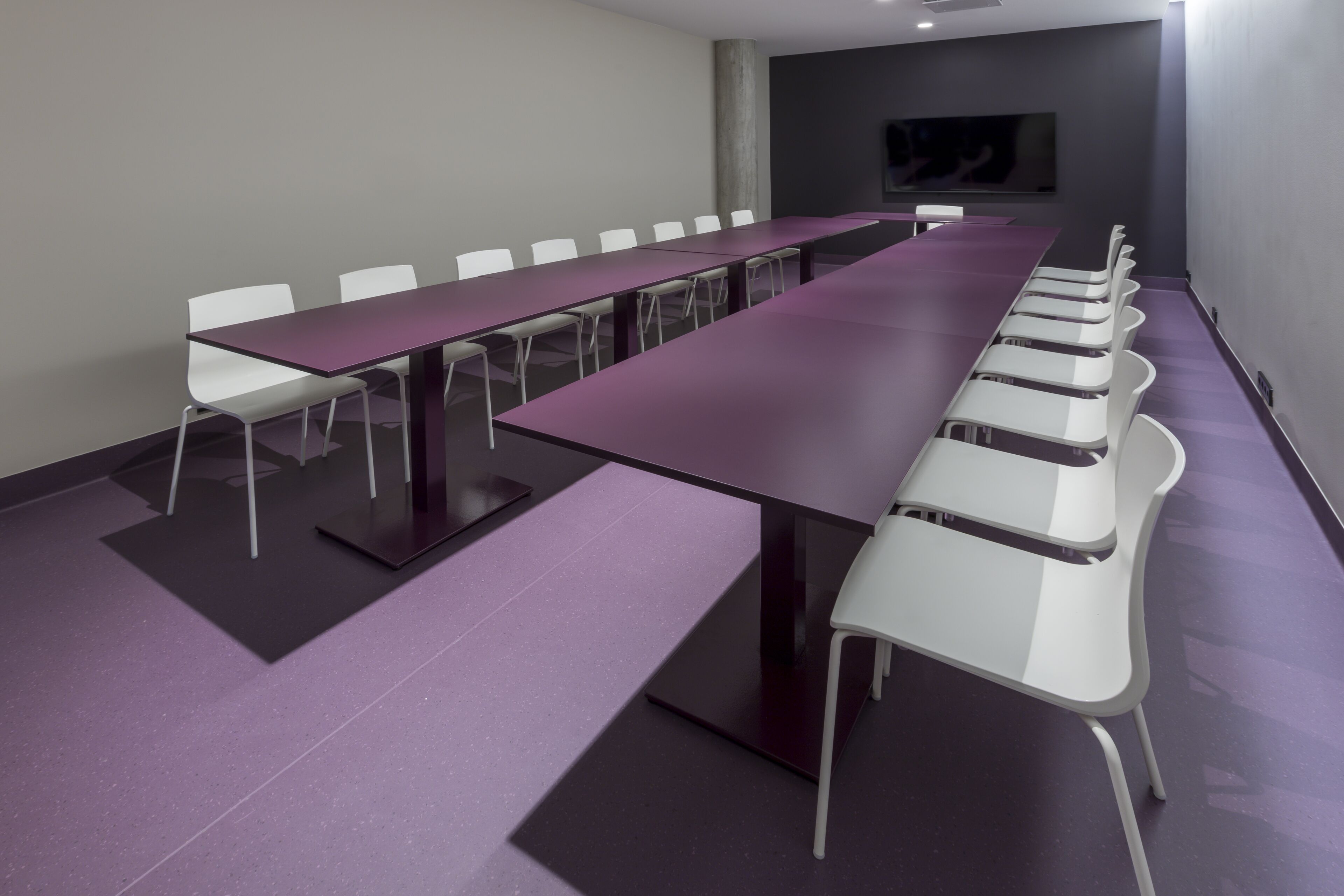 Ein Konferenzraum mit einem langen Tisch, weißen Stühlen, einer großen Säule und einem Flachbildfernseher an der Wand.