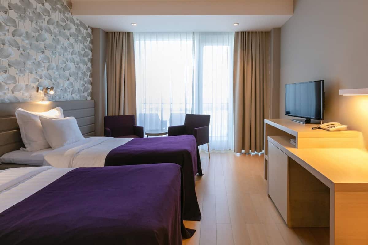A hotel room with two beds, a desk with a TV and phone, and a view of the city through large windows.