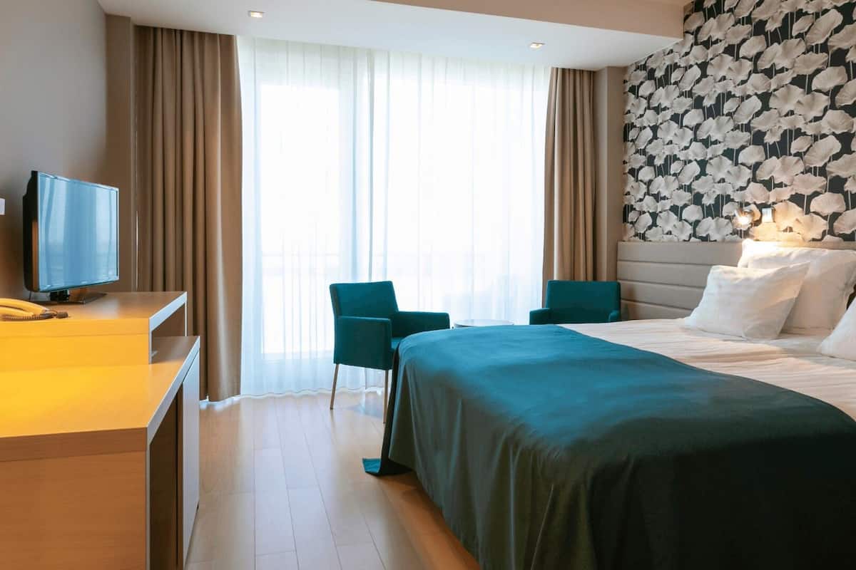 A hotel room with a bed, a desk with a telephone, two chairs, and a large window with curtains.