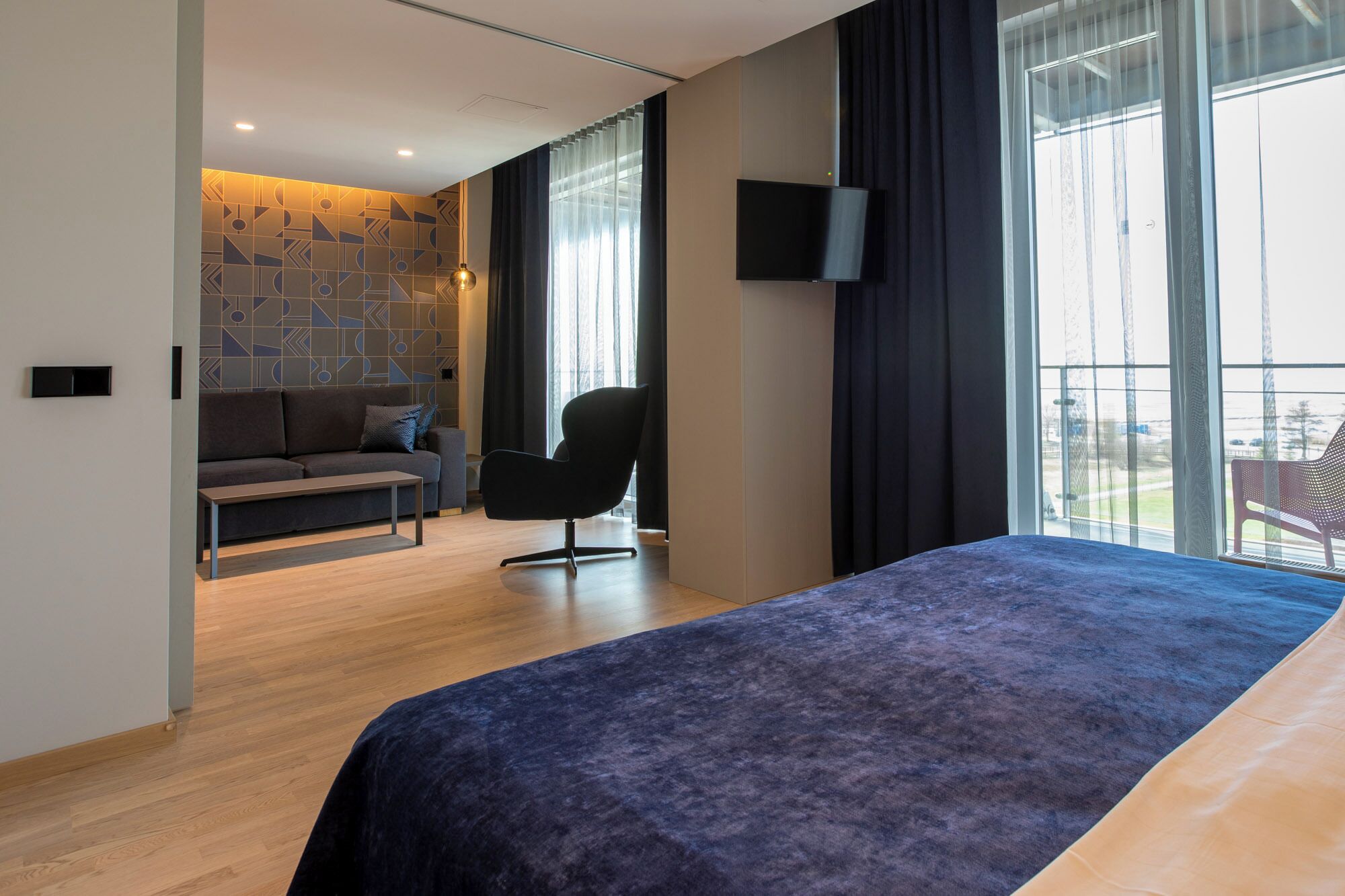 Una camera d'albergo moderna con un letto, una poltrona, una televisione e ampie finestre.