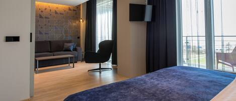 A modern hotel room with a bed, a sofa, a chair, a TV, and large windows.