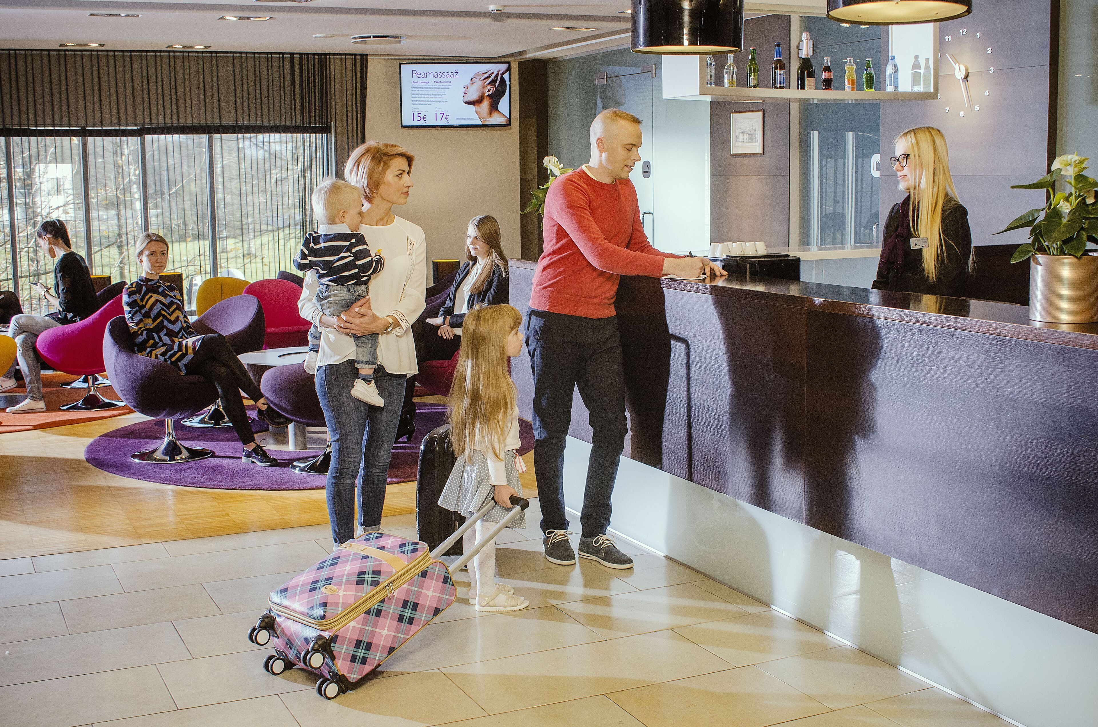 Un área de recepción de hotel con diseño moderno, que incluye un mostrador de recepción, zona de estar con sillas coloridas y un amplio ventanal que proporciona luz natural.