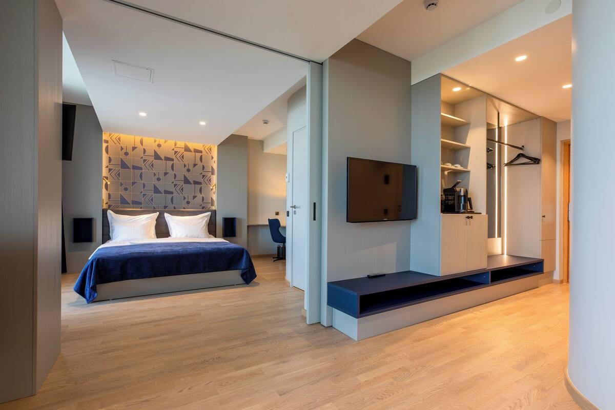 A modern hotel room with a large bed, a built-in TV, a walk-in closet, and a geometric-patterned headboard.