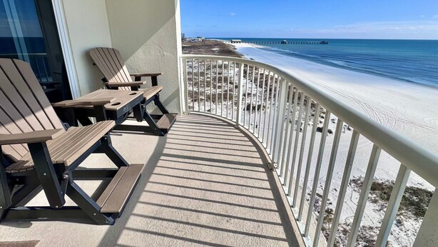 A balcony with two Adirondack chairs and a table, overlooking a beach with a pier. - Time to get… 🎶 Knee deep in the water somewhere! 🎶 on island time! (Gulf Shores)