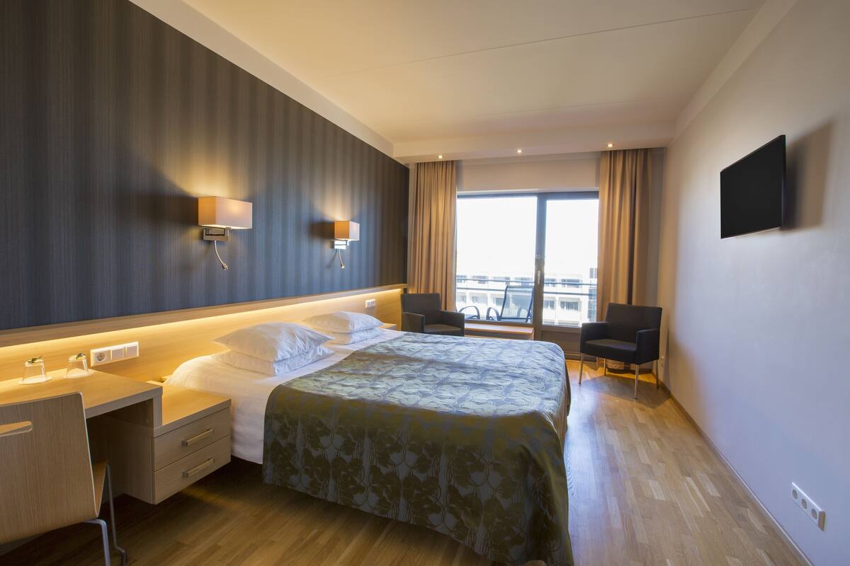 A modern hotel room with a large bed, a desk, and a chair. There is a TV mounted on the wall and a window with curtains.