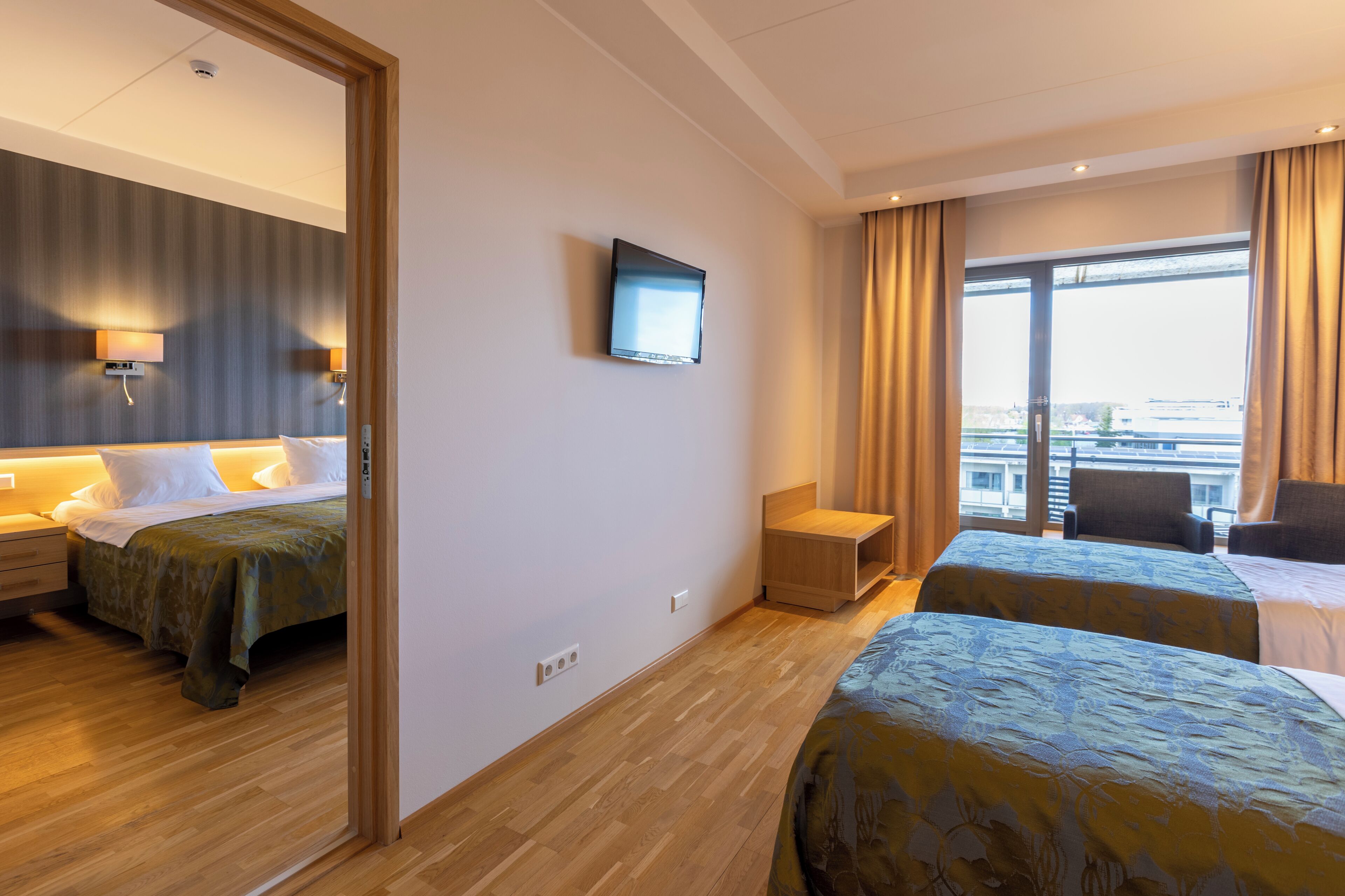 Habitación de hotel con dos camas, un televisor, una mesita y vistas a la ciudad a través de amplios ventanales.