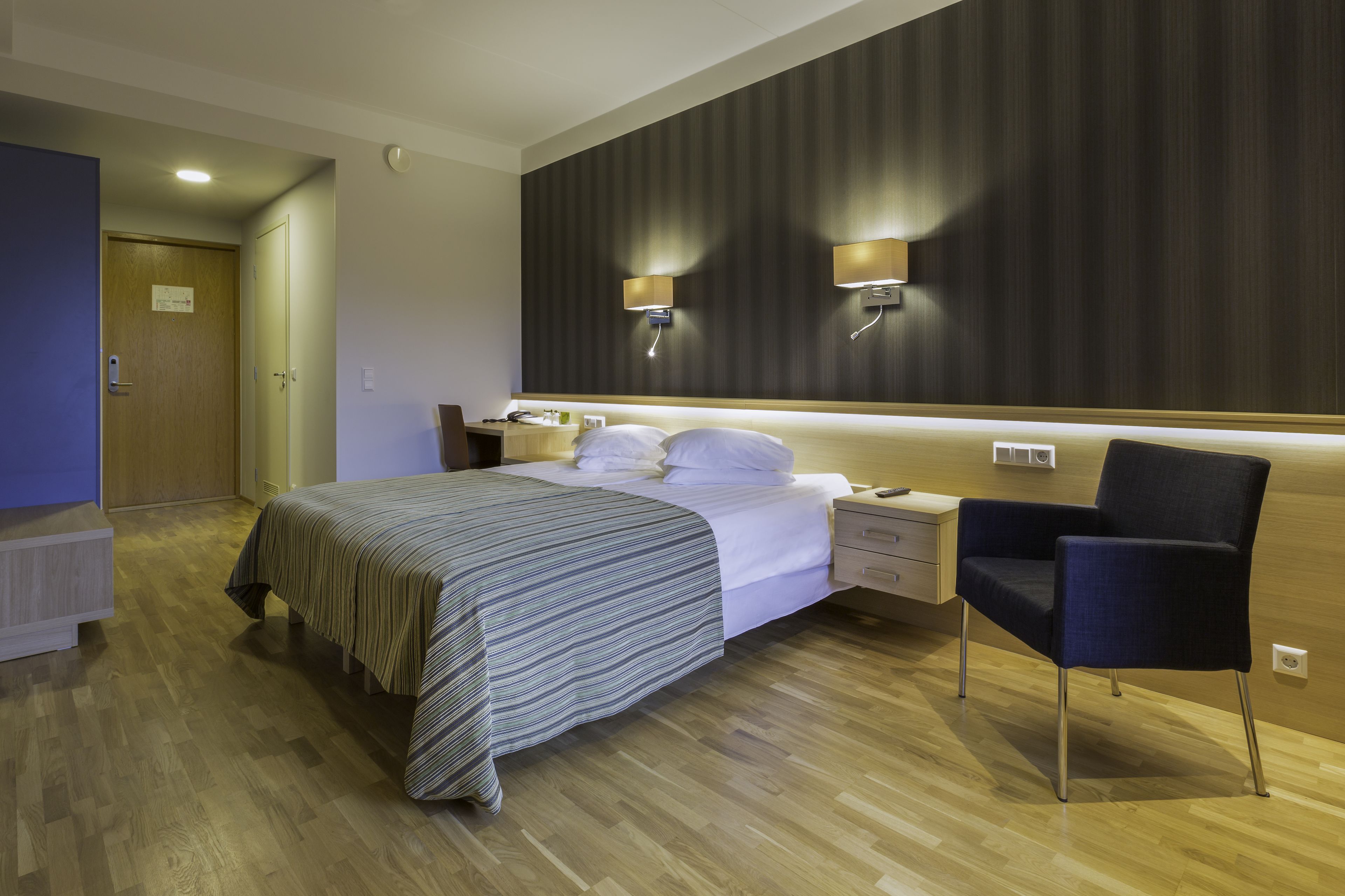 A hotel room with a bed, bedside tables, a chair, and a wall-mounted lamp.