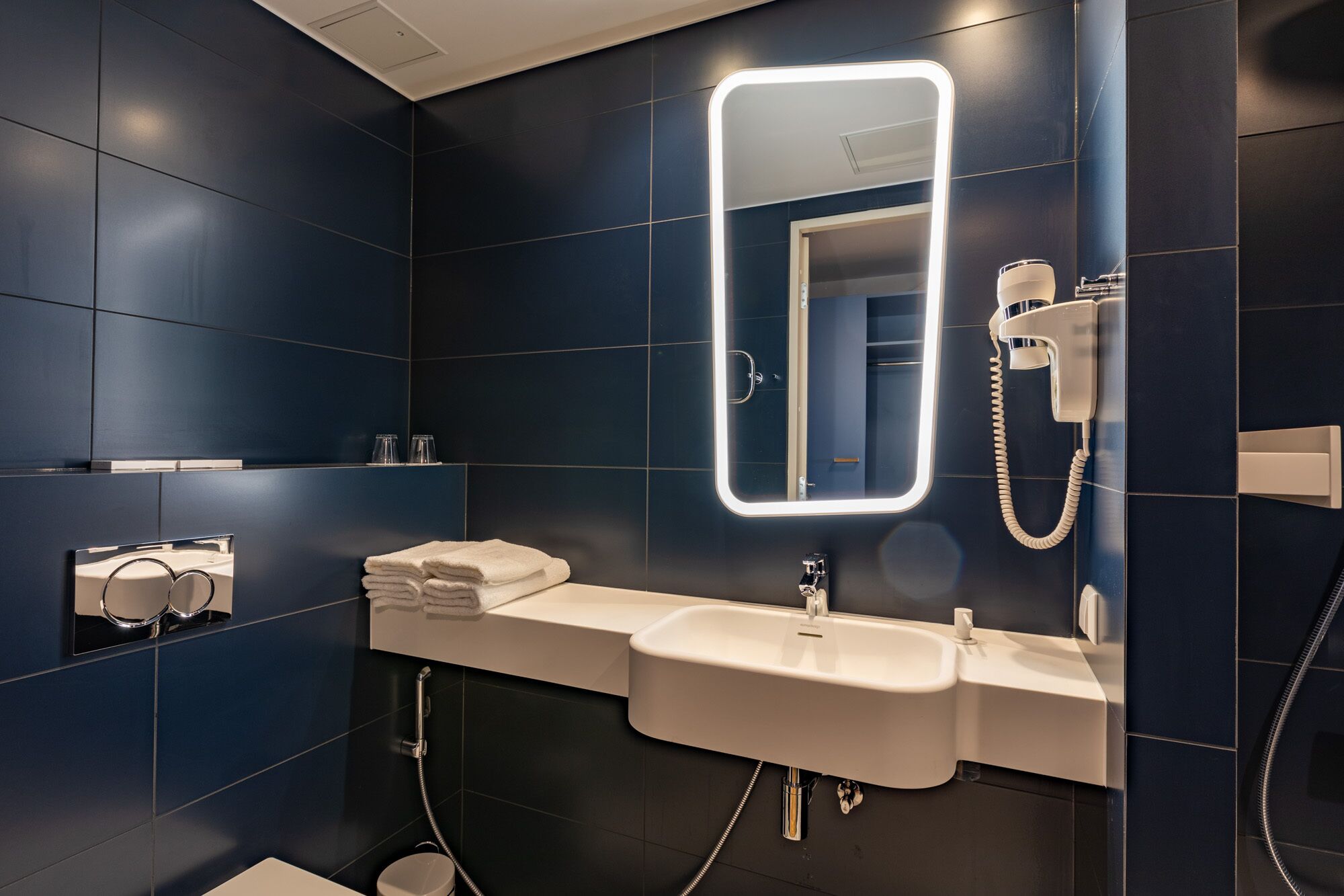 A modern bathroom with dark blue tiles, a large mirror, a white sink, and a hairdryer.