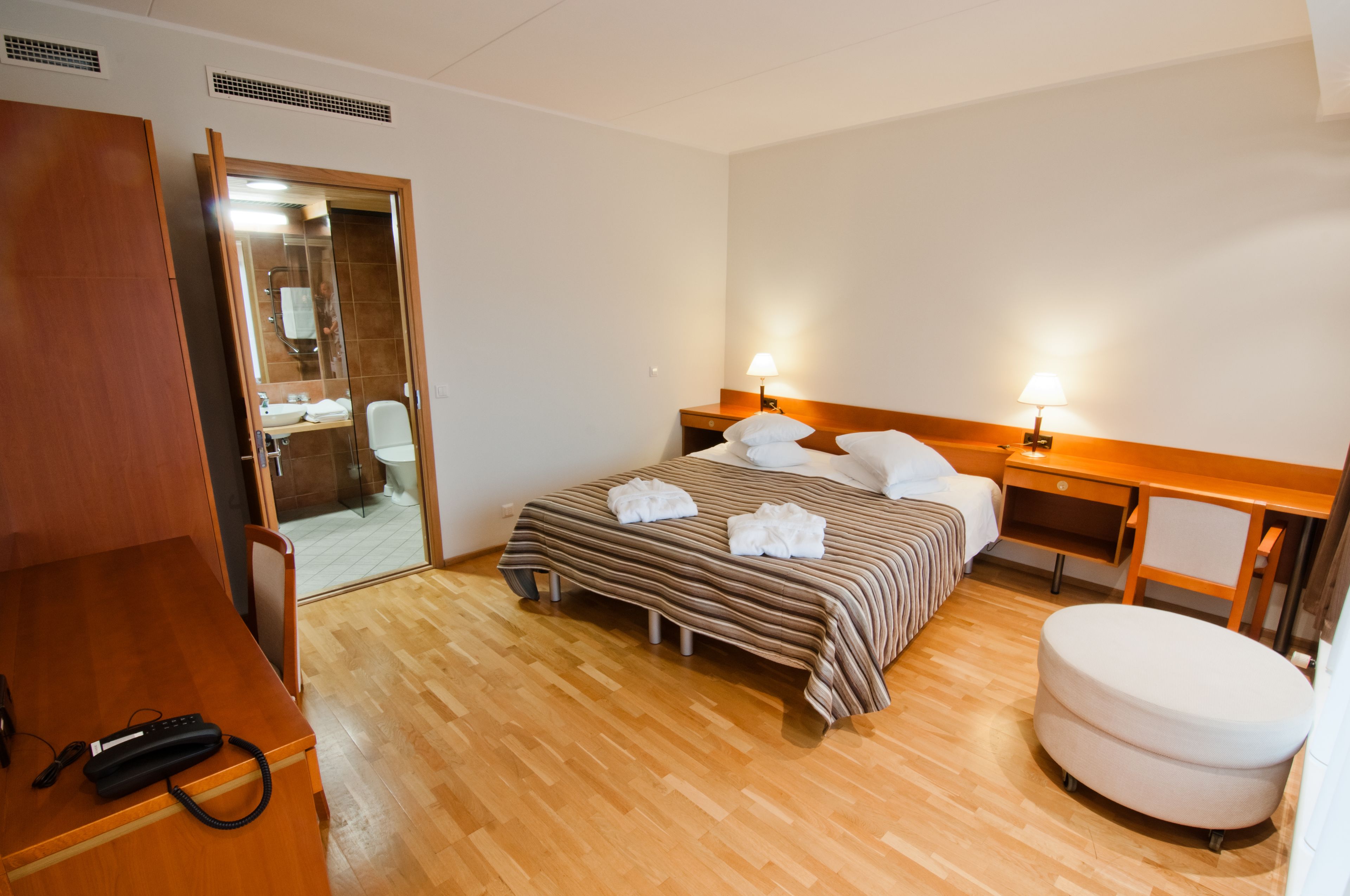 A hotel room with a wooden floor, a bed with striped bedding, two bedside lamps, a wooden nightstand, a white round ottoman, and a wooden wardrobe.